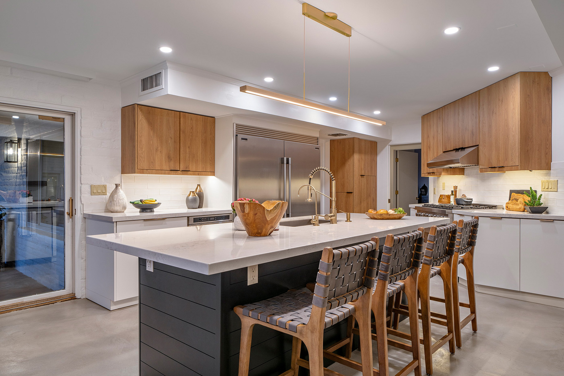 Contemporary kitchen featuring a large waterfall-edge quartz island with woven barstools, custom wood cabinetry, stainless steel appliances, brass fixtures, white countertops, and warm recessed lighting.