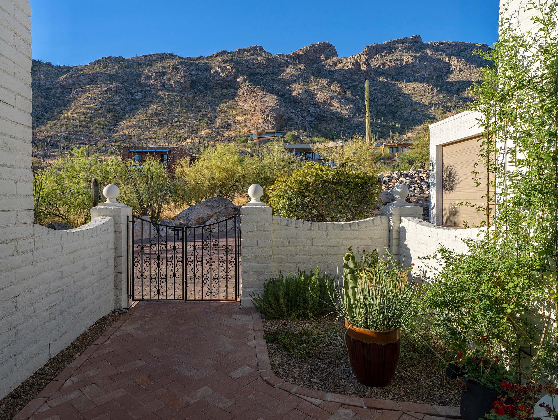 Brick-paver entry courtyard framed by white masonry walls, wrought-iron gate, desert landscaping, and dramatic mountain views in the background.