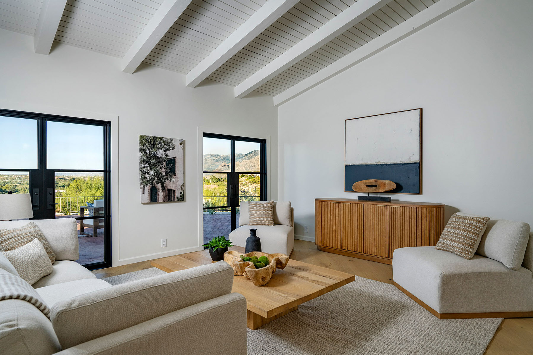 Bright contemporary great room featuring vaulted beam ceilings, custom furnishings, natural wood accents, large black-framed windows, and views of the surrounding mountains and outdoor living spaces.