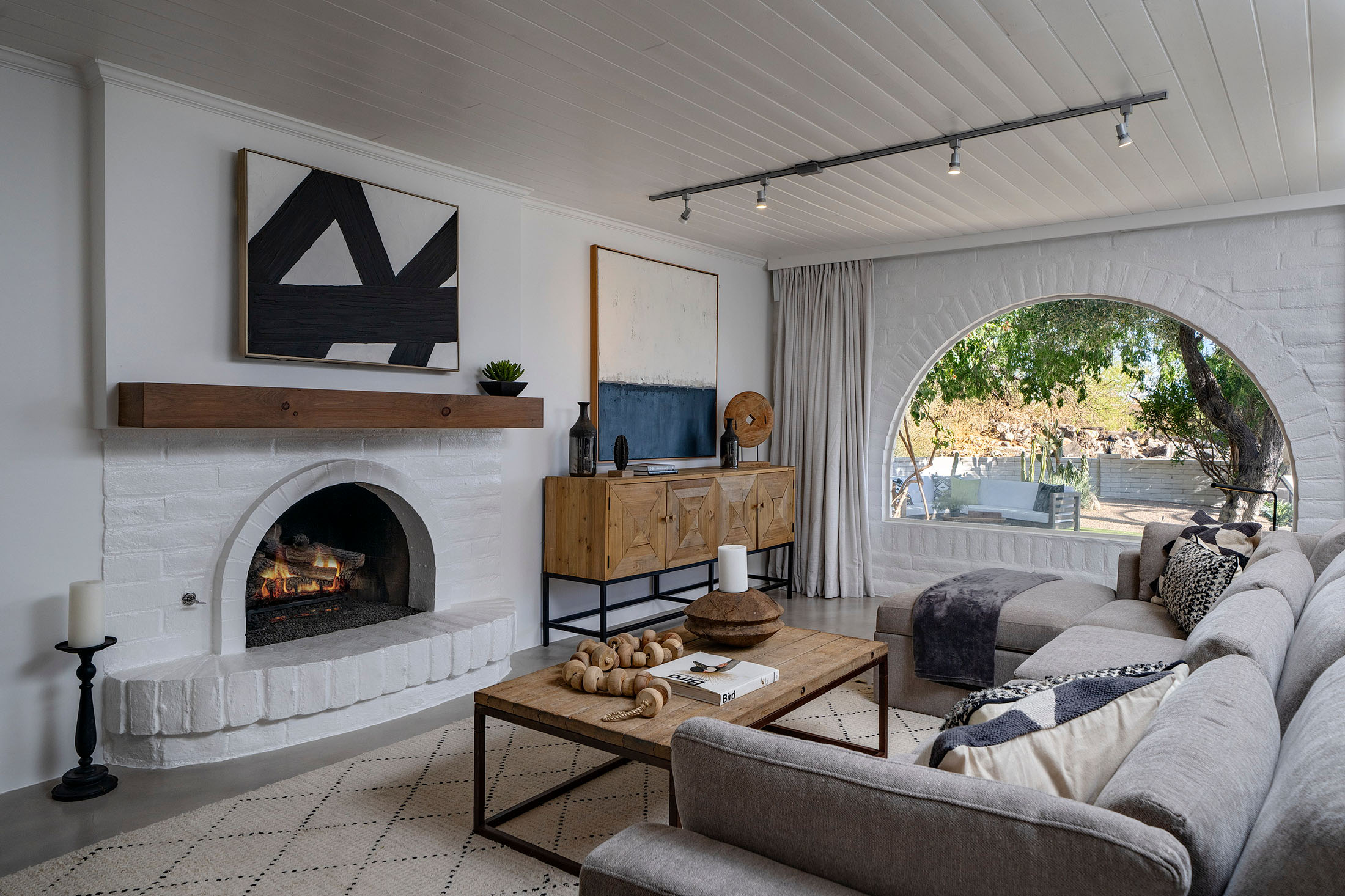 Sophisticated living room featuring a white-painted brick fireplace, large arched picture window, neutral furnishings, contemporary artwork, and natural light.