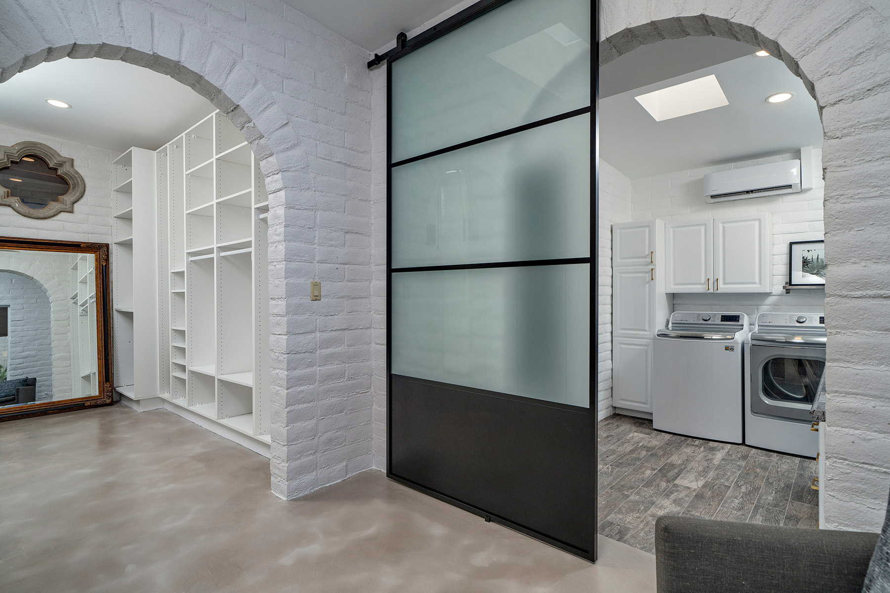 Spacious walk-in closet with custom built-in shelving, polished concrete flooring, arched openings, frosted-glass sliding door, and adjoining laundry room with storage cabinetry.
