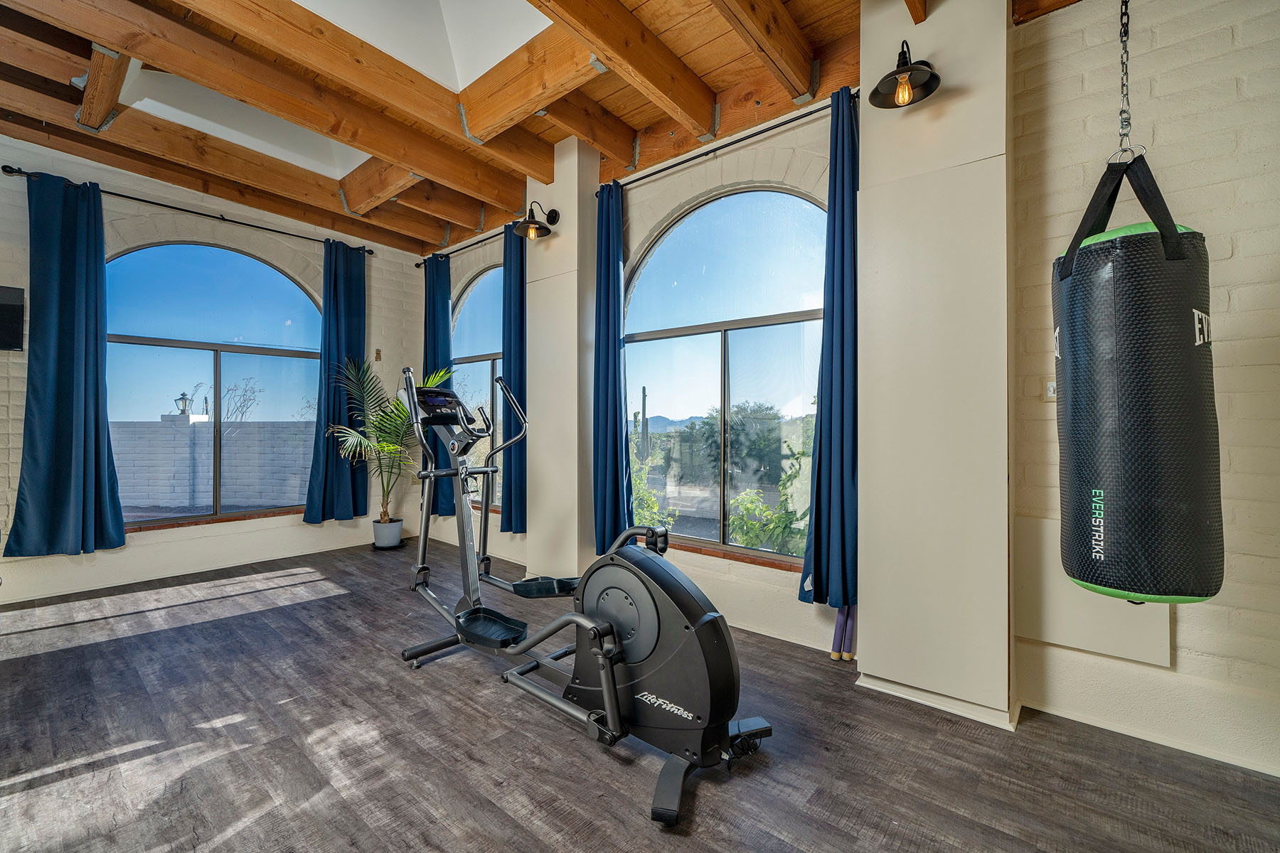 Spacious home gym featuring large arched windows, exposed wood-beam ceiling, cardio equipment, punching bag, and abundant natural light.