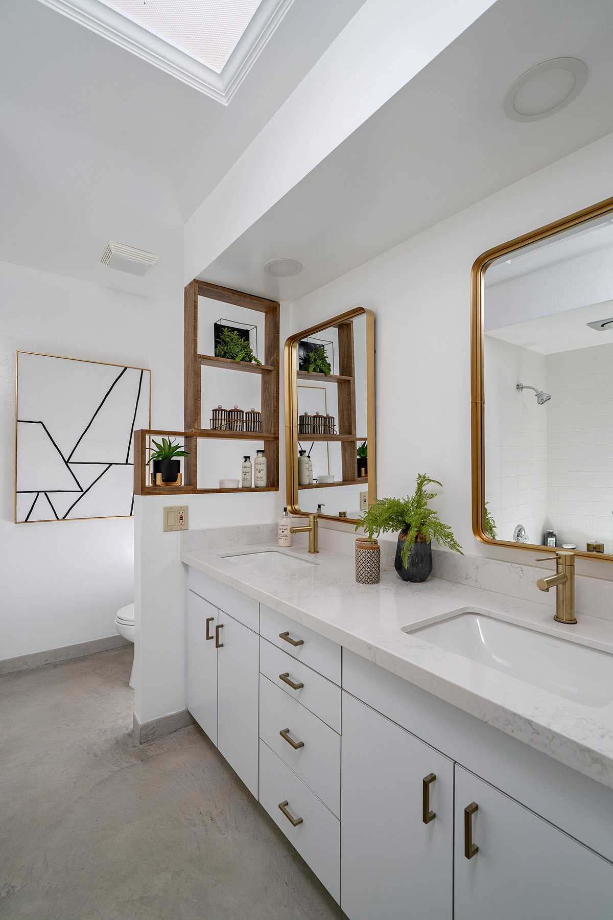 Modern primary bathroom featuring dual sinks, quartz countertops, brass fixtures, skylight, custom shelving, and bright contemporary finishes